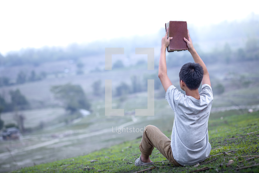 a boy sitting on a hill holding a Bible 