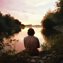 Young woman sitting on the bank of the river and looking at the sunset