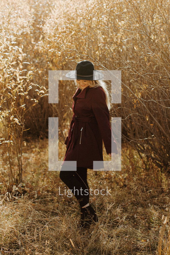 a young woman in a hat and coat standing outdoors in golden sunlight 