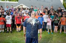 A youth pastor, covered in colorful cake and frosting, facing a wild and rowdy crowd of youth group students in the back.