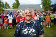 A youth pastor smiling wildly, covered in colorful cake and frosting, in front of a rowdy youth group in the background.