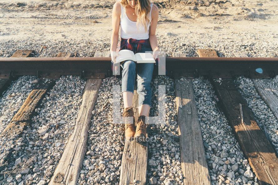 A young girl sitting on railroad tracks reading her bible.