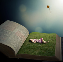 woman lying in green grass on the pages of a Bible flying a kite 