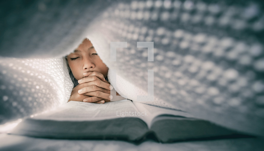 a little boy reading a Bible and praying under a blanket