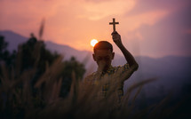 a boy holding a cross at sunset praying in a field 