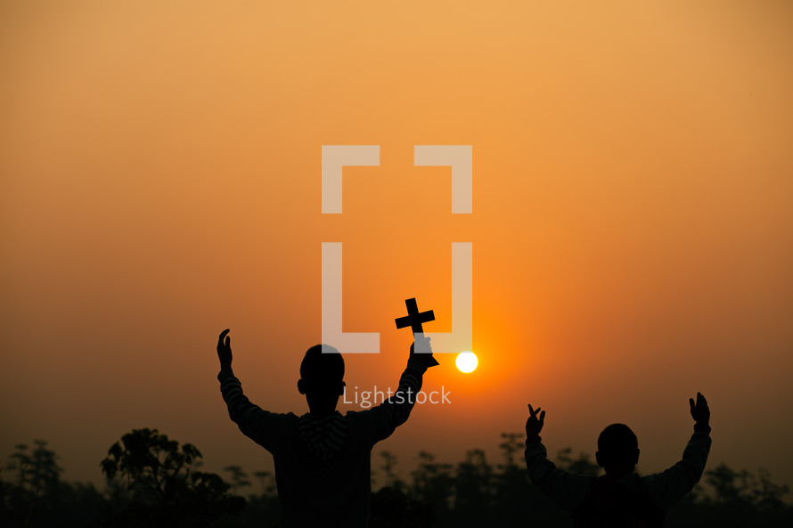 two boys holding up a cross at sunset under an orange sky 