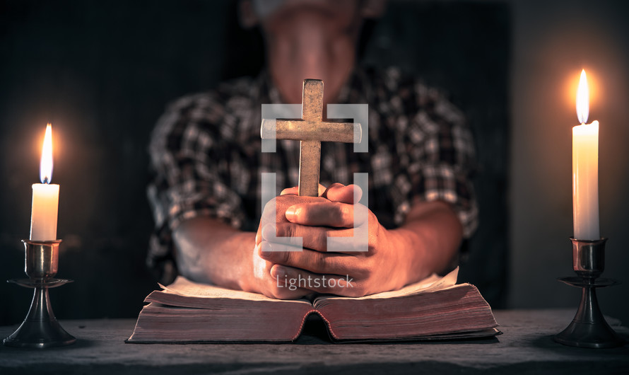 a man holding a cross and praying over a Bible in candlelight 