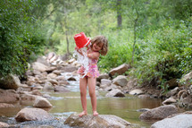 a girl child playing with a bucket of water in a stream 