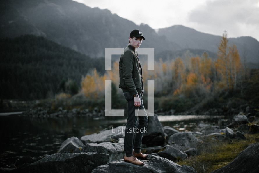 a young man in dress shoes standing on rocks by a lake 