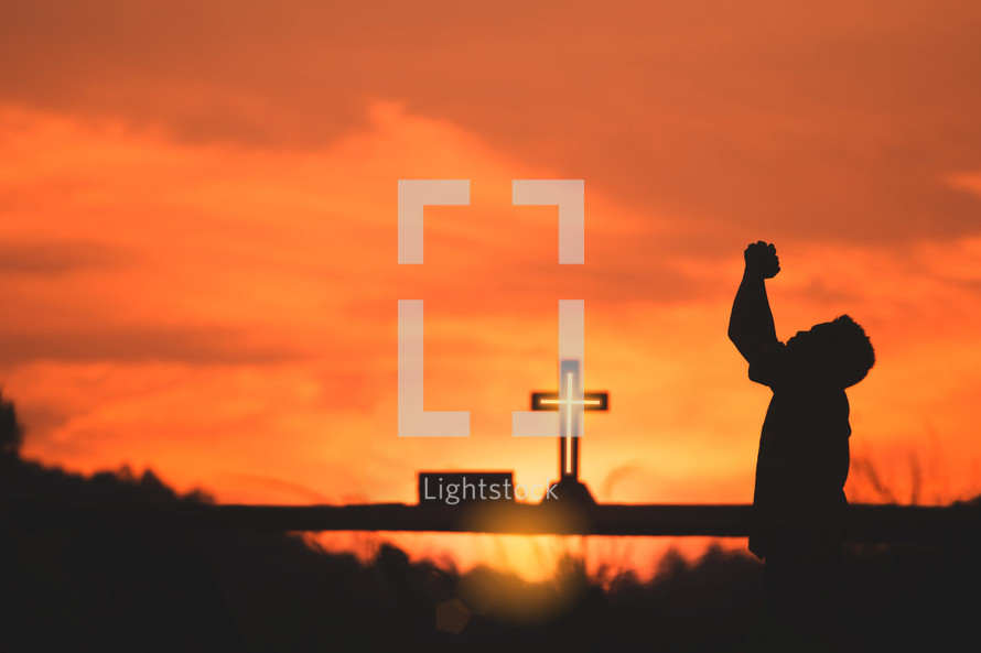 A boy praying to God with a cross at sunset 