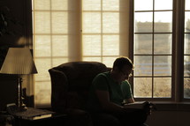 Man sitting in a chair by a window, reading the Bible.