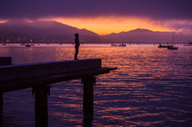 silhouette of a boy standing at the end of a dock under a purple sunset 