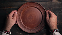 Ukrainian man sitting in national restaurant and waiting for order. Guy knocks fingers nervously on wooden table while waiting for dish. Empty clay bowl with spoon.