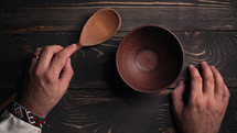Ukrainian man sitting in national restaurant and waiting for order. Guy knocks fingers nervously on wooden table while waiting for dish. Empty clay bowl with spoon.