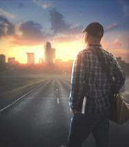 A man walks on a road towards a city with a bag and a Bible
