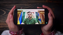 August 2022 - Kyiv, Ukraine. Watching online speech of President Volodymyr Zelenskiy on smartphone on wooden table background. News from front. russian aggression.
