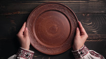 Female hands with empty ukrainian clay plate from wooden table. Woman in traditional embroidered shirt. Top view. National tableware with handmade ornament
