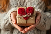 Caring hands present a handmade gift wrapped in brown paper and tied with twine, topped with a knitted red heart, symbolizing love and thoughtfulness