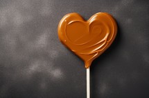 Heart-shaped caramel candy on a stick against a dark textured background