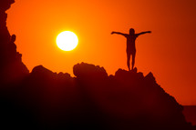 silhouette of a woman standing on a mountain with outstretched arms at sunset 