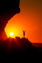 silhouette of a woman standing on a mountain with raised hands at sunset 