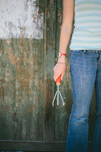 Teen holding a garden claw tool standing near a half painted fence.