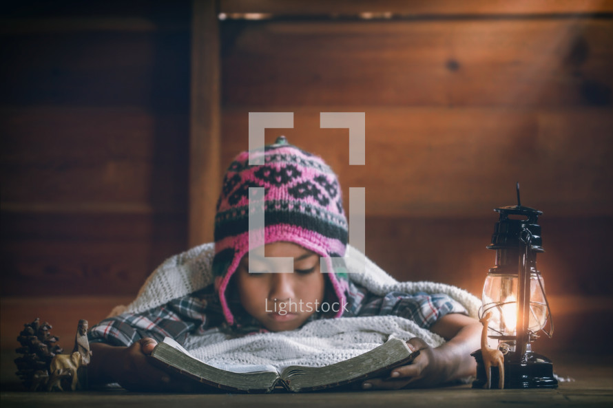 a child in a beanie reading a Bible under a blanket by a lantern 