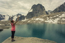 a woman with raised hands standing by the edge of a lake surrounded by snow covered mountains 
