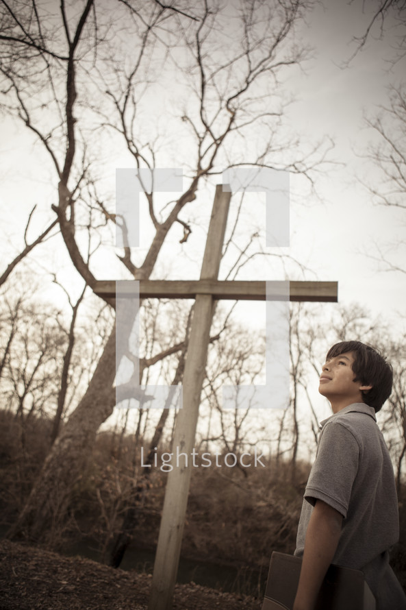 teen boy standing near a cross holding a Bible outdoors 