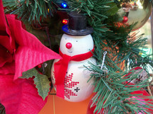 A snowman Christmas Tree ornament sits on the branch of a Christmas tree surrounded by red leaves and other beautiful Christmas ornament decorations in the celebration of Christmas time.  Frosty the Snowman was a favorite Christmas character we grew up with as kids and he has found a home here among familiar friends that adorn and decorate this Christmas tree to bring in the  Christmas holiday season. 