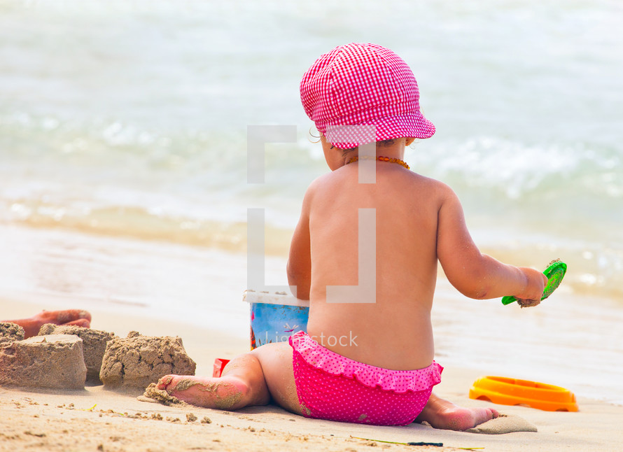 Little girl plays on the shoreline at the beach wearing a hat.