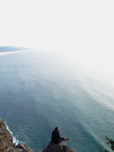 Man sitting on a rock on the edge of a cliff overlooking the ocean.
