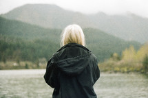 A blonde woman stands before a mountain lake.