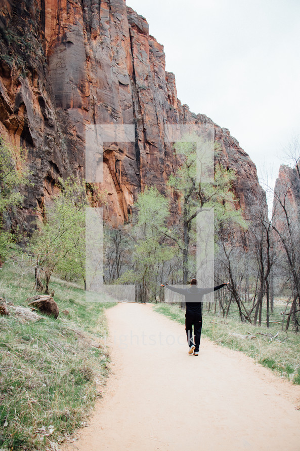 man walking along a paved path and red rock cliffs 