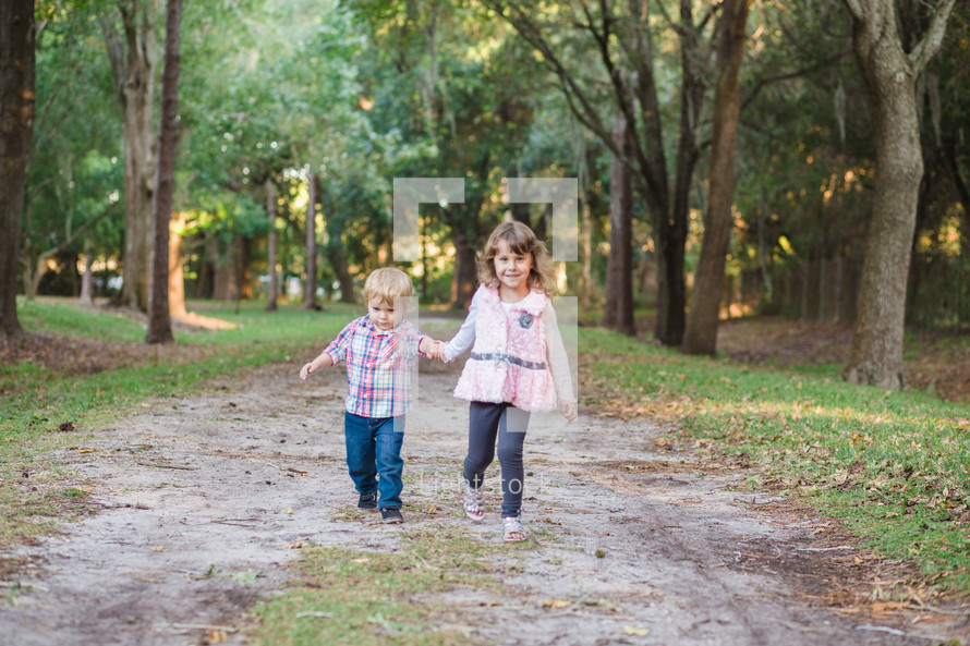 brother and sister holding hands walking on a dirt road 