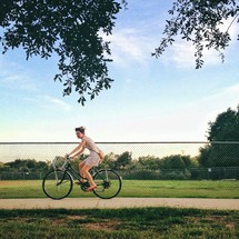 Woman riding bike on sidewalk through park with chain link fence and trees.