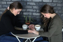 friends sitting at a table with coffee mugs holding hands in prayer 