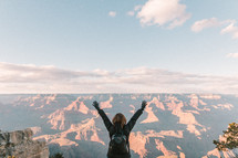 a woman standing at the edge of a cliff in front of canyons with arms raised 
