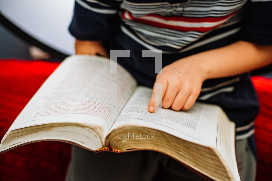 A young boy reads an open Bible while pointing to the words with his pointer finger.