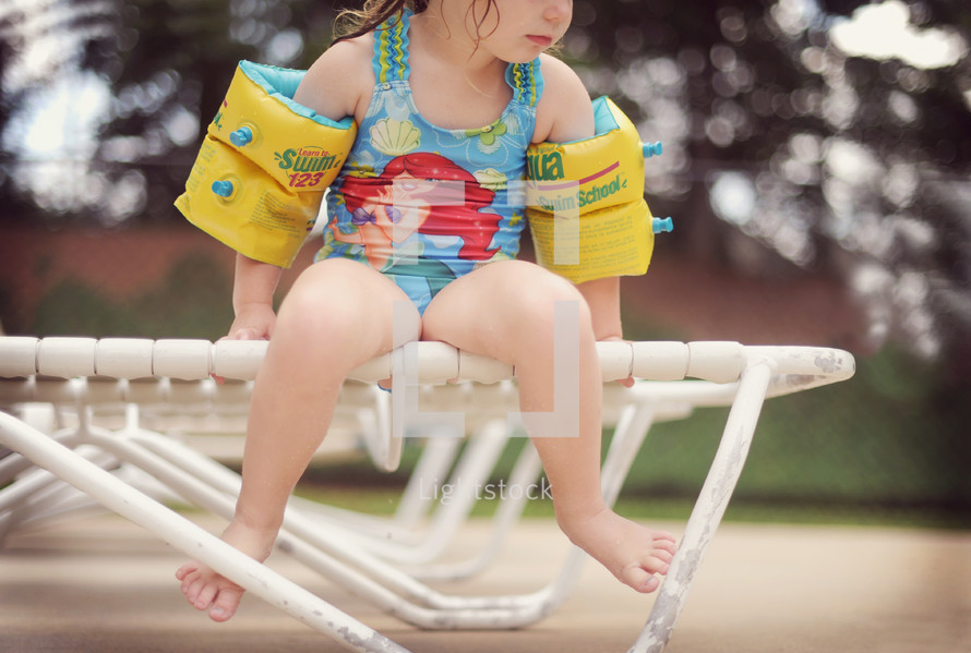 a toddler sitting on a lounge chair at a pool in a swim suit and floaties