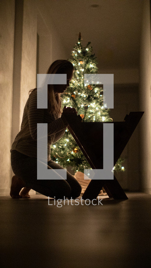 a woman kneeling to pickup a baby out of a manger in front of a Christmas tree 