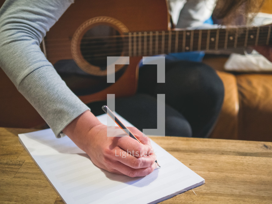 A woman sits on a couch with a guitar while writing on music notation  paper