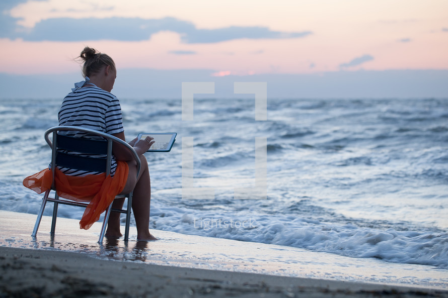 Woman sitting on chair by sea and using ipad
