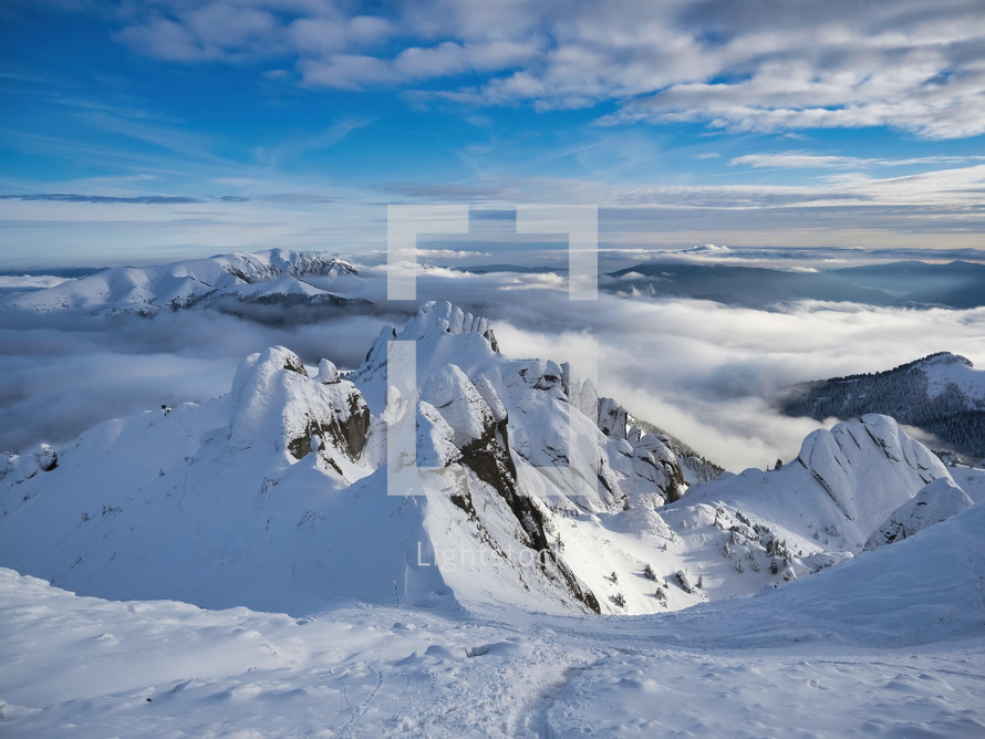 Aerial view of distant mountain peaks above clouds with a clear, sunny, blue sky.
