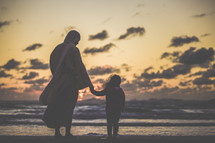 Jesus holding hands with a little girl standing on a beach at sunset 