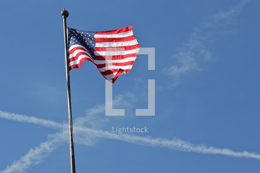 American flag on a flag pole with airplane vapor trails in the sky in the shape of a cross