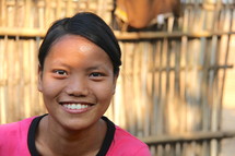 A smiling Asian girl with a bamboo fence in the background.