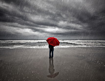 man with a red umbrella standing on a beach in the rain 
