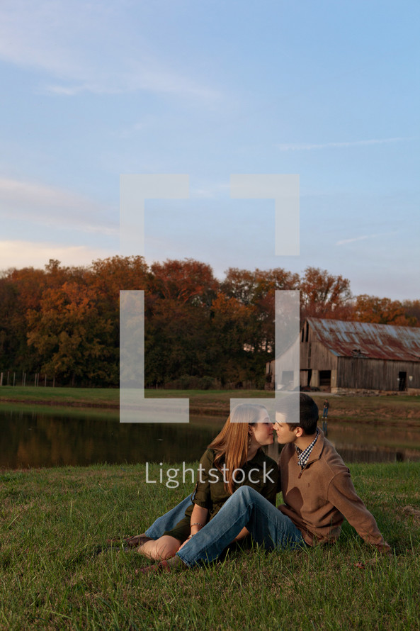 Man and woman kissing while sitting on grassy meadow by pond with barn in the background.