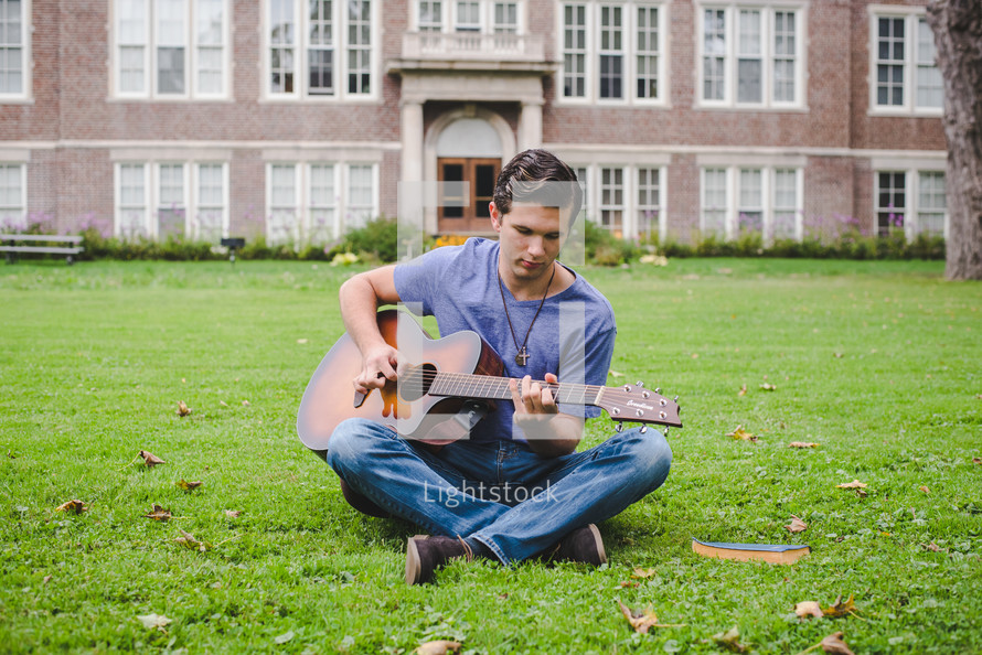 A young man sitting in grass playing a guitar 